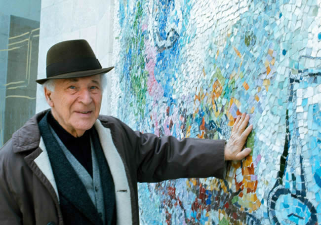 Artist Marc Chagall wearing a hat presses his hand against a wall with blue mosaic designs.