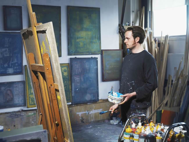 Male artist holding paint brush looking at canvas on easel in studio