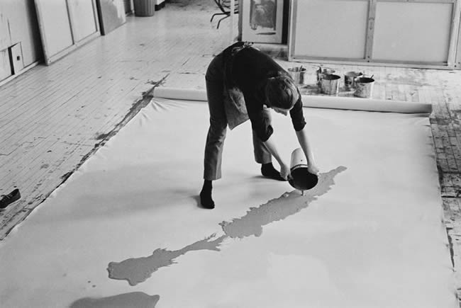 Helen Frankenthaler pouring paint carefully onto a canvas on the floor.