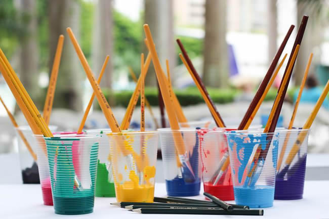 Different paint colors in cups with art brushes sticking out.