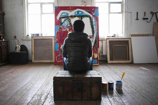 Male artist sitting on crate, looking at artwork, rear view