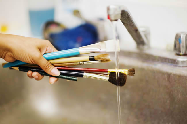 Person's hand washing paint brushes under a faucet